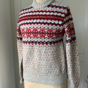 May include: A gray turtleneck sweater with a patterned design in red, navy, and yellow. The sweater has a ribbed collar, cuffs, and hem. The body of the sweater features a repeating pattern of small red dots.