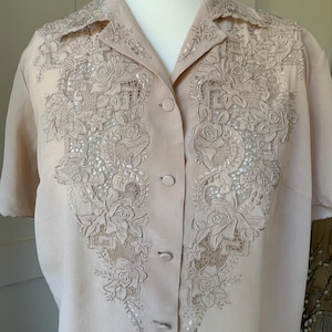 May include: A short-sleeved, light beige button-down shirt featuring intricate tonal embroidery and lace panels on the front. The design includes detailed floral motifs, specifically roses, integrated with geometric patterns along the collar and chest.