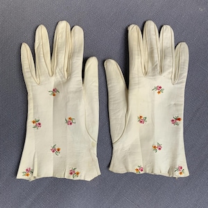 May include: A pair of off-white leather gloves featuring small, scattered floral embroidery in shades of pink, yellow, and green. The gloves have a smooth finish and a slightly flared wrist opening, with the floral pattern distributed across the back.