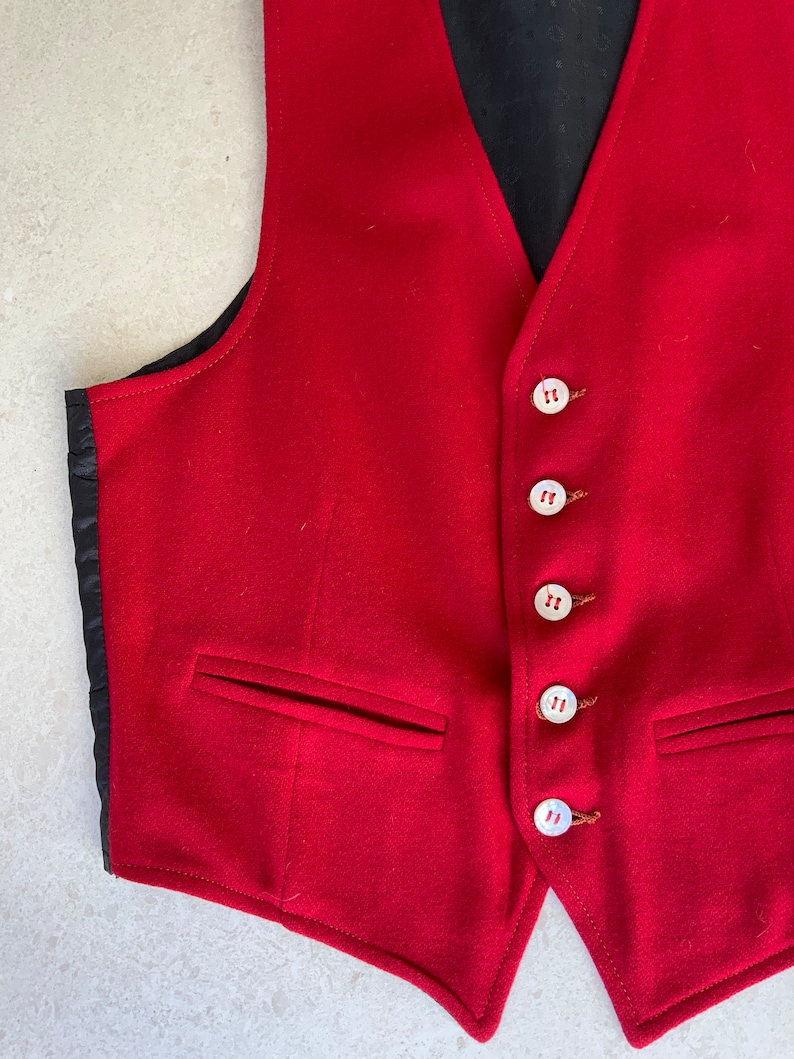 K&ouml;nnte beinhalten: Eine rote Wollweste mit sechs wei&szlig;en Kn&ouml;pfen und zwei Taschen. Die Weste hat ein schwarzes Futter.
