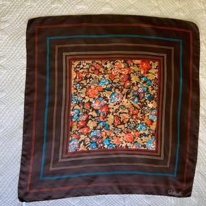 K&ouml;nnte beinhalten: Ein quadratisches braunes Tuch mit einem Blumenmuster in der Mitte. Das Blumenmuster umfasst orange, blaue und wei&szlig;e Blumen auf einem beigen Hintergrund. Das Tuch hat einen braunen Rand mit roten und blauen Streifen.
