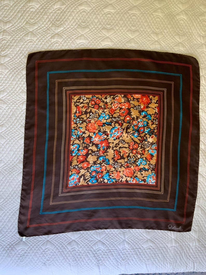 K&ouml;nnte beinhalten: Ein quadratisches braunes Tuch mit einem Blumenmuster in der Mitte. Das Blumenmuster umfasst orange, blaue und wei&szlig;e Blumen auf einem beigen Hintergrund. Das Tuch hat einen braunen Rand mit roten und blauen Streifen.