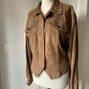 K&ouml;nnte beinhalten: Eine braune Wildlederjacke mit Kragen, langen &Auml;rmeln und zwei Pattentaschen auf der Vorderseite. Die Jacke hat eine leicht verk&uuml;rzte L&auml;nge und einen spitzen Saum.