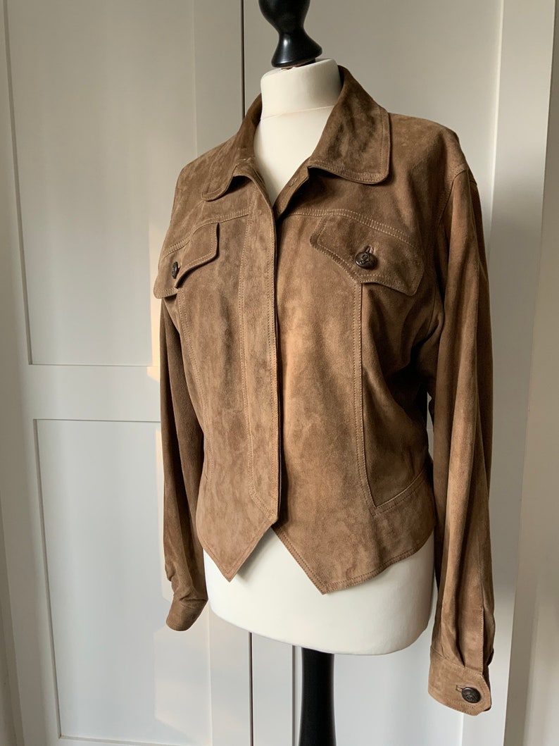 K&ouml;nnte beinhalten: Eine braune Wildlederjacke mit Kragen, langen &Auml;rmeln und zwei Pattentaschen auf der Vorderseite. Die Jacke hat eine leicht verk&uuml;rzte L&auml;nge und einen spitzen Saum.