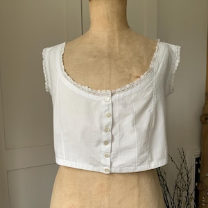 May include: A white, sleeveless top with a scoop neckline and a row of buttons down the front. The neckline and armholes are trimmed with delicate lace. The garment is displayed on a mannequin.