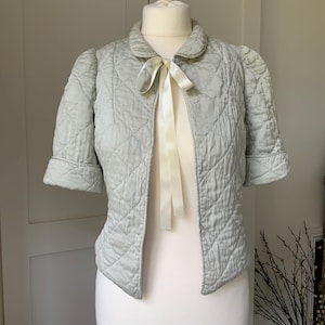May include: A light blue quilted jacket with short sleeves and a cream-colored ribbon tie. The jacket has a diamond pattern and a rounded collar. The jacket is displayed on a white mannequin.