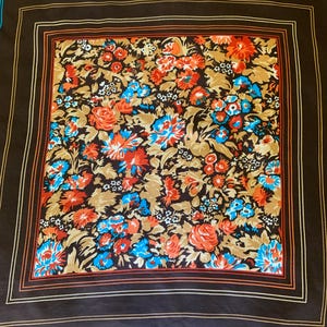 K&ouml;nnte beinhalten: Ein quadratisches Tuch mit einem Blumenmuster in Orange, Blau und Wei&szlig; auf braunem Grund. Der Schal hat einen dunkelbraunen Rand mit orangefarbenen und blauen Streifen.
