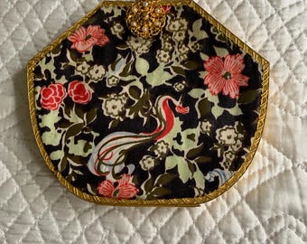 Könnte beinhalten: Eine achteckige Clutch mit goldfarbenem Rahmen und Verschluss. Der Stoff zeigt ein Blumenmuster mit rosa und roten Blüten, grünen Blättern und einem stilisierten Vogel auf schwarzem Hintergrund. Der Verschluss ist mit kleinen Perlen verziert.