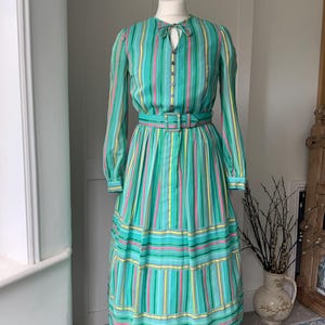 May include: A vintage long dress with vertical stripes in teal, pink, and yellow. The dress has long sleeves, a belted waist, and a tiered skirt. It features a keyhole neckline and button details. The dress is a classic design.