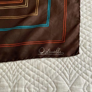 K&ouml;nnte beinhalten: Braunes Seidentuch mit floralem Design in der Ecke. Der Schal hat einen quadratischen Rand mit orangefarbenen und blauen Linien. Der Markenname "Ostivelli" ist kursiv in der Ecke geschrieben.