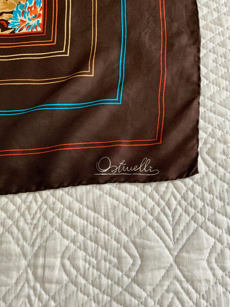 K&ouml;nnte beinhalten: Braunes Seidentuch mit floralem Design in der Ecke. Der Schal hat einen quadratischen Rand mit orangefarbenen und blauen Linien. Der Markenname "Ostivelli" ist kursiv in der Ecke geschrieben.
