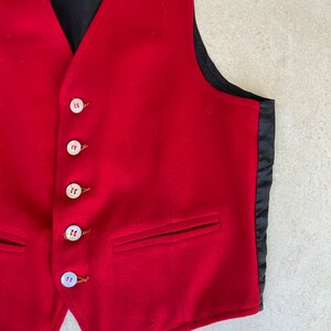 K&ouml;nnte beinhalten: Eine rote Wollweste mit schwarzem Satin-Futter und sechs wei&szlig;en Kn&ouml;pfen. Die Weste hat zwei Taschen auf der Vorderseite.