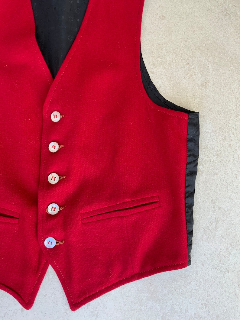 K&ouml;nnte beinhalten: Eine rote Wollweste mit schwarzem Satin-Futter und sechs wei&szlig;en Kn&ouml;pfen. Die Weste hat zwei Taschen auf der Vorderseite.