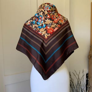 May include: A square scarf with a brown background and a floral pattern in shades of red, blue, and tan. The scarf has brown, red, and blue stripes along the edges. The scarf is displayed on a white mannequin.