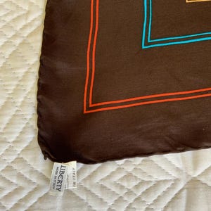 K&ouml;nnte beinhalten: Ein braunes Seidentuch mit geometrischem Design mit orangefarbenen, blauen und beigefarbenen R&auml;ndern. Das Tuch hat ein Etikett mit der Aufschrift "LIBERTY Made in Italy."