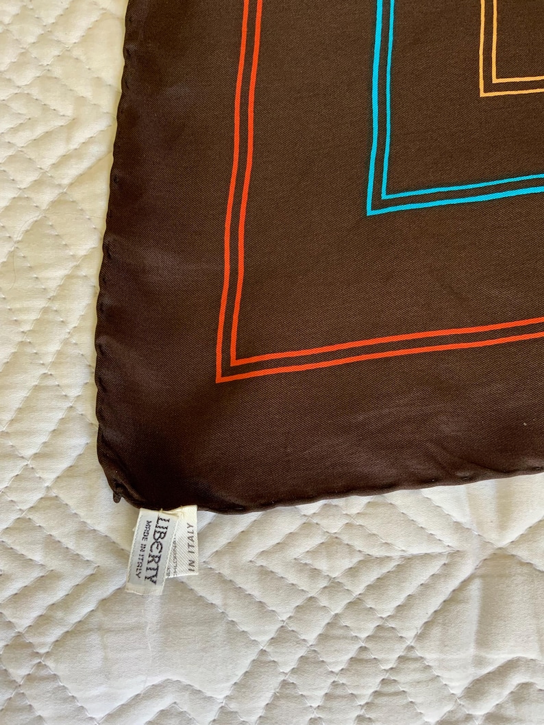 K&ouml;nnte beinhalten: Ein braunes Seidentuch mit geometrischem Design mit orangefarbenen, blauen und beigefarbenen R&auml;ndern. Das Tuch hat ein Etikett mit der Aufschrift "LIBERTY Made in Italy."