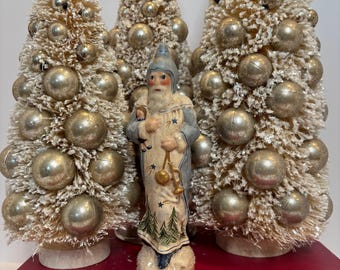 May include: Three bottle brush trees with silver ornaments and a central figure of a bearded person in a blue hooded coat holding a small doll and a gold trumpet, standing on a red book titled Works William Shakespeare.