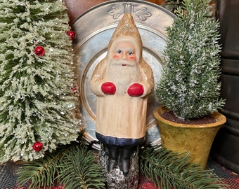 May include: A white and red Santa Claus figurine with a glittery beard and hat, standing on a wooden log. The figurine is holding red mittens and is surrounded by small artificial pine trees.