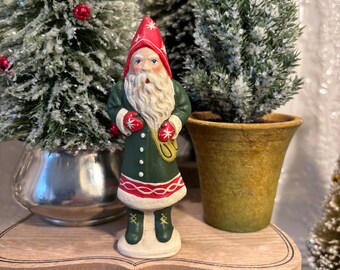 May include: A ceramic Santa Claus figurine with a red hat and mittens, green coat, and white beard. The figurine is standing on a wooden shelf with small Christmas trees in the background. The Santa is holding a gold sack.