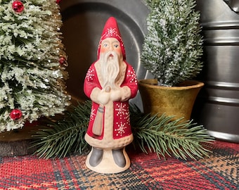 May include: A small, red and white Santa Claus figurine with a white beard and a red hat. The figurine is standing on a plaid surface with two small, snow-covered Christmas trees in the background.