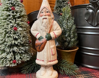 May include: A ceramic Santa Claus figurine in a white coat with red trim and green gloves. The figurine has a long white beard and is holding a bag. Two small Christmas trees and a plaid tablecloth are in the background.
