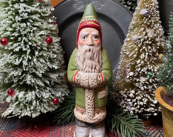 May include: A ceramic Santa Claus figurine with a green coat, red and gold hat, and white beard. The figurine is standing in front of several small Christmas trees. The background includes a metal plate and a red and black plaid tablecloth.