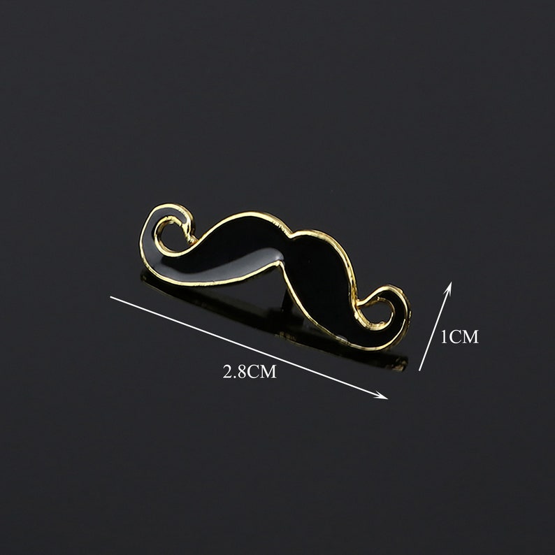 K&ouml;nnte beinhalten: Eine schwarze und goldene Schnurrbartnadel mit goldener Umrandung. Die Nadel ist 2,8 cm breit und 1 cm hoch.