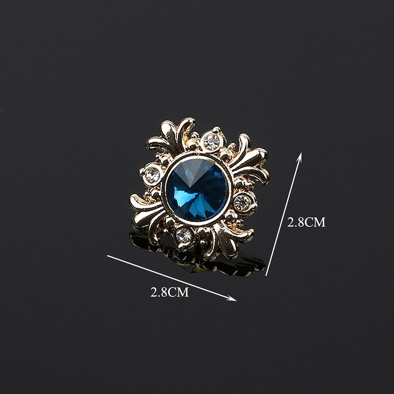 K&ouml;nnte beinhalten: Eine goldfarbene dekorative Brosche mit einem blauen Strassstein in der Mitte, umgeben von kleineren klaren Strasssteinen. Die Brosche hat einen Durchmesser von 2,8 cm.