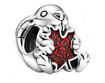 Puede incluir: Un charm de plata con un conejo sosteniendo una hoja de arce roja. El conejo tiene la nariz y los ojos negros. El charm está diseñado para usarse en una pulsera o collar.