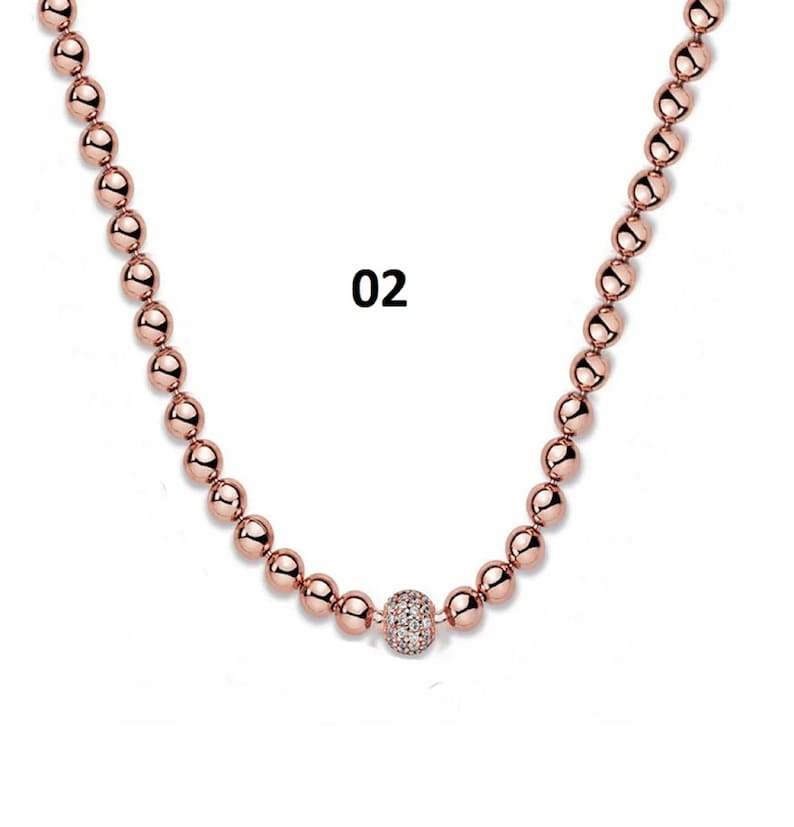 May include: A rose gold necklace with a single larger bead in the center that is encrusted with clear rhinestones. The necklace is made of small round beads.