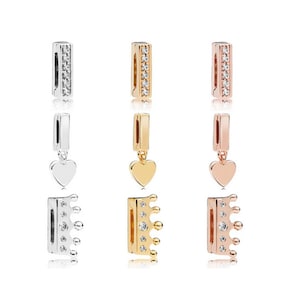 Op de afbeelding: Drie metalen bedelhangers in zilver, goud en roségoud. Elke bedel heeft een rechthoekige staaf met ingebedde heldere stenen. De bedels bevatten een hartvormige hanger en een kroonhanger, met een verscheidenheid aan sieradenontwerpen.