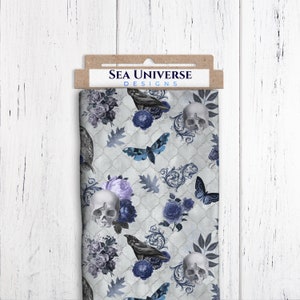 Könnte beinhalten: Ein weißer Stoff mit einem sich wiederholenden Muster aus blauen Rosen, Schädeln, Schmetterlingen, Raben und Blättern. Der Stoff hat einen abgenutzten, Vintage-Look. Der Text "SEA UNIVERSE DESIGNS" ist auf einem braunen Etikett gedruckt, das am Stoff befestigt ist.