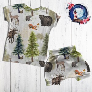 May include: A white children's clothing set with a woodland animal print. The set includes a short-sleeved shirt and shorts. The print features bears, moose, deer, raccoons, foxes, and trees.