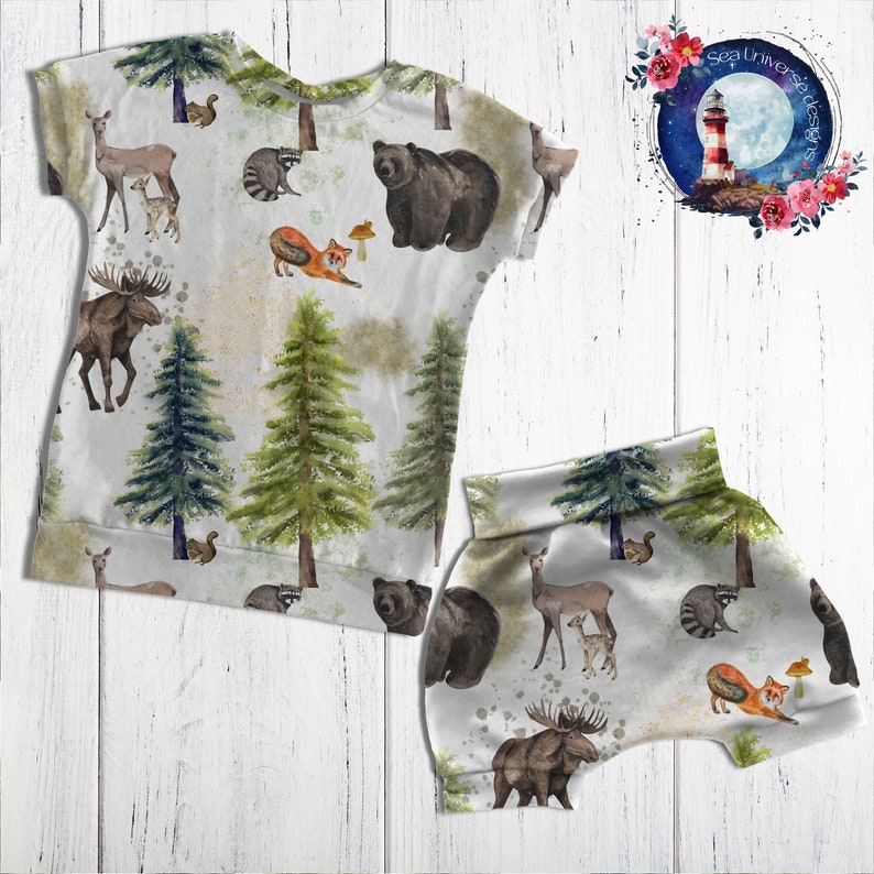 May include: A white children's clothing set with a woodland animal print. The set includes a short-sleeved shirt and shorts. The print features bears, moose, deer, raccoons, foxes, and trees.