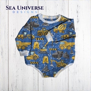 Peut inclure: Une grenouillère bleue pour bébé sur le thème de la construction. La grenouillère présente un motif répétitif de véhicules de chantier jaunes, notamment un camion à benne basculante, un bulldozer, une excavatrice et une bétonnière. L'arrière-plan est un motif de briques bleues avec du ruban adhésif de sécurité noir et jaune.
