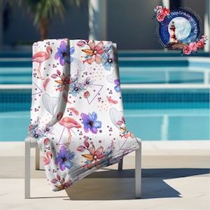 May include: A white beach towel with a pink flamingo, blue and pink flowers, and gray geometric shapes. The towel is folded and placed on a white lounge chair by a swimming pool.