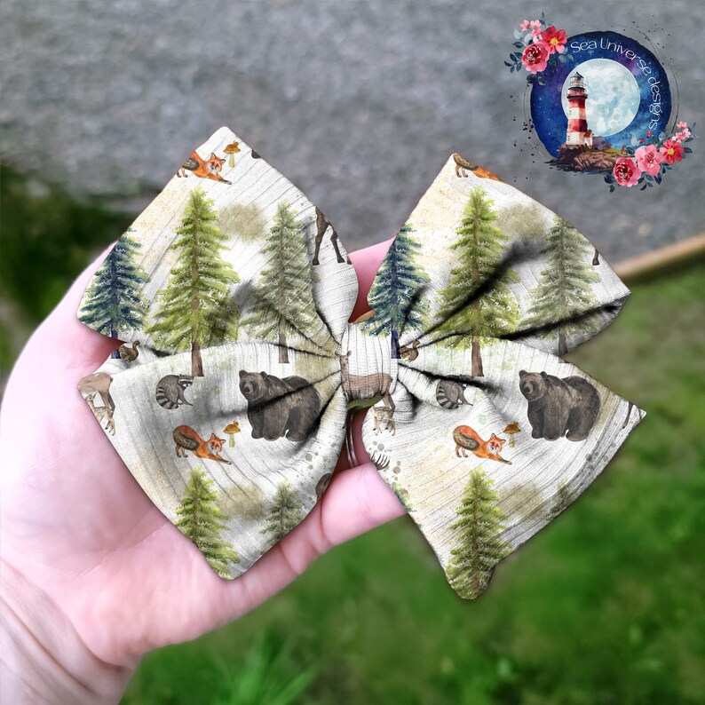 May include: A white fabric bow with a woodland theme. The bow features a repeating pattern of brown bears, foxes, and pine trees on a white background.