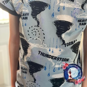 Peut inclure: Un t-shirt bleu avec un imprimé blanc et noir de tornades, de nuages, d'éclairs et de gouttes de pluie. Le texte "Heavy Rain", "Tornado" et "Thunderstorm" est imprimé sur le t-shirt.
