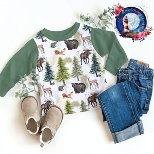 May include: A white long-sleeve shirt with a green raglan sleeve design. The shirt features a woodland animal print with moose, bears, deer, raccoons, and trees. The shirt is laid out on a white surface with a pair of brown boots and blue jeans.