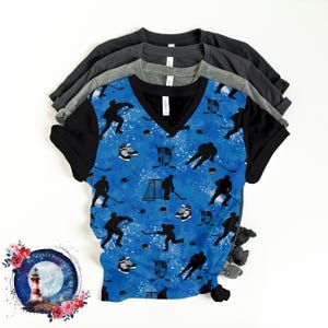 May include: A blue t-shirt with a black v-neck collar and a hockey-themed pattern. The pattern features black silhouettes of hockey players, pucks, and hockey sticks on a blue background with white speckles.