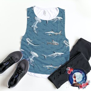 Könnte beinhalten: Ein blaues ärmelloses Tanktop mit weißem Besatz und einem Muster aus Haien und Hammerhaien. Das Tanktop liegt auf einer weißen Oberfläche mit einem Paar schwarzer Turnschuhe und schwarzen Leggings.