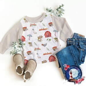 May include: A beige long-sleeved shirt with a white raglan sleeve and a white background with a repeating pattern of red barns, pumpkins, ducks, and baskets of apples. The shirt is laid out on a white surface with a pair of brown boots and blue jeans.