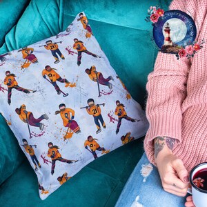 May include: A white throw pillow with a repeating pattern of hockey players in various poses. The players are wearing orange, yellow, and purple jerseys with numbers and stars. The background is a light blue with white splatters of paint.