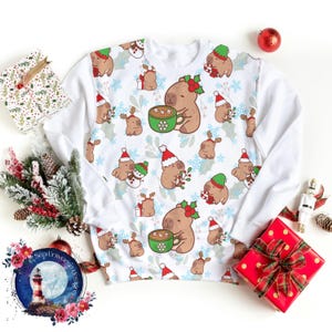 May include: White sweatshirt with a repeating pattern of cartoon capybaras, reindeer, and snowflakes. Some capybaras wear Santa hats and hold mugs of hot chocolate. The sweatshirt has long white sleeves and is surrounded by Christmas decorations.