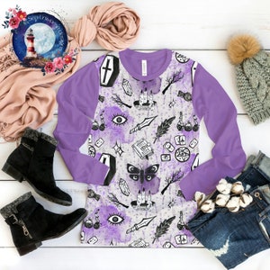 May include: A long-sleeved shirt with a purple and white design. The shirt features a variety of witchy symbols, including a cauldron, a book, a knife, and a feather. The shirt is perfect for anyone who loves Halloween or witchcraft.