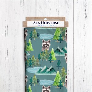 May include: Sea Universe Designs fabric featuring a repeating pattern of raccoons, pine trees, and mountains on a teal blue background.