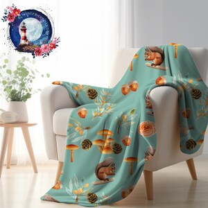May include: A teal blue fleece throw blanket with a repeating pattern of squirrels, mushrooms, acorns, and pine cones. The blanket is draped over a white armchair.