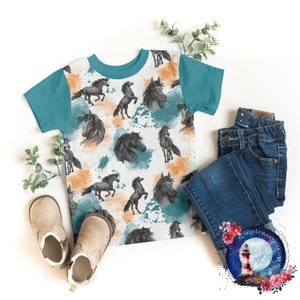 May include: A white t-shirt with a teal blue raglan sleeve design. The shirt features a repeating pattern of black horses with watercolour splashes in shades of orange and teal blue.