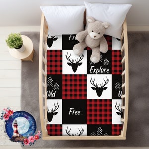 May include: A black and white cot sheet with a red and black buffalo check pattern. The sheet features a repeated design of a deer head with antlers, the words "Free", "Wild", and "Explore", and a stylised pine tree design.
