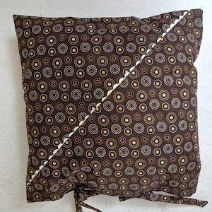 Könnte beinhalten: Ein braunes Kissen mit einem weißen und braunen Kreis-Muster. Das Kissen hat einen weißen Zierstich entlang der diagonalen Naht und Bänder an der Unterseite.
